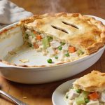 Chicken pot pie in a white oval dish with a golden puff pastry lid, one portion served out showing a thick filling of chicken, carrot, celery and peas