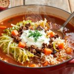 Unstuffed cabbage roll soup with beef and rice in a red Dutch oven, topped with sour cream and fresh dill