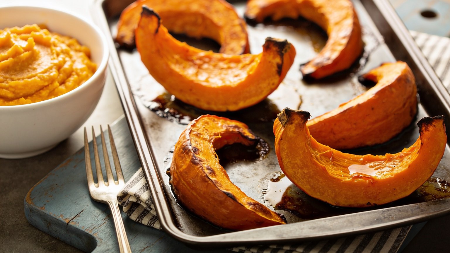 roasted pumpkin wedges in their skin on a dark metal baking tray beside a white bowl of mashed pumpkin, a fork and a striped napkin on a blue-grey board