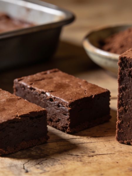 Three fudgy cocoa powder brownies on weathered wood, one on end with a dense cut face and cocoa-dusted top, the cut slab in its pan and a cocoa dish behind