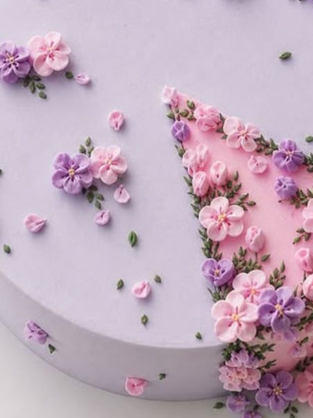 Lilac buttercream flower cake on a white board on marble, pink and purple piped flowers crowded around a pink frosted wedge set flush in the lilac top