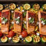 Sheet pan salmon and zucchini — four paprika-crusted fillets in a dark pan with zucchini rounds, whole cherry tomatoes, corn, crumbled feta and basil
