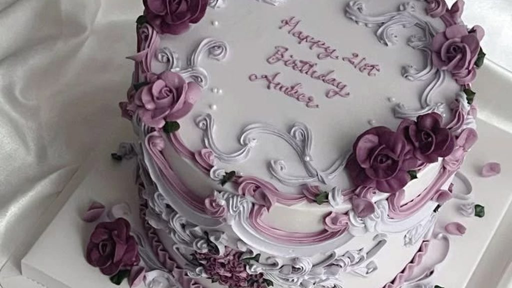 Vintage buttercream cake piped with white and pink scrollwork, draped swags and deep plum roses, with a birthday message written across the ivory top