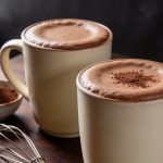 Healthy hot chocolate in two cream stoneware mugs with a fine cocoa-dusted foam, a bowl of cocoa powder and a wire whisk on a walnut table.
